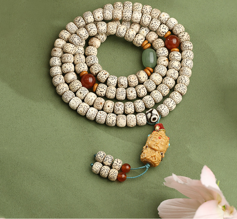 Cream speckled meditation beads coiled on a muted green surface, with red and green accents, a sculpted pendant and two beaded tails.