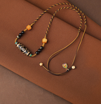 Inner Compass necklace on rust-colored fabric, showing its elongated eye-pattern bead, braided cord, pale tail beads and multicolor charm.