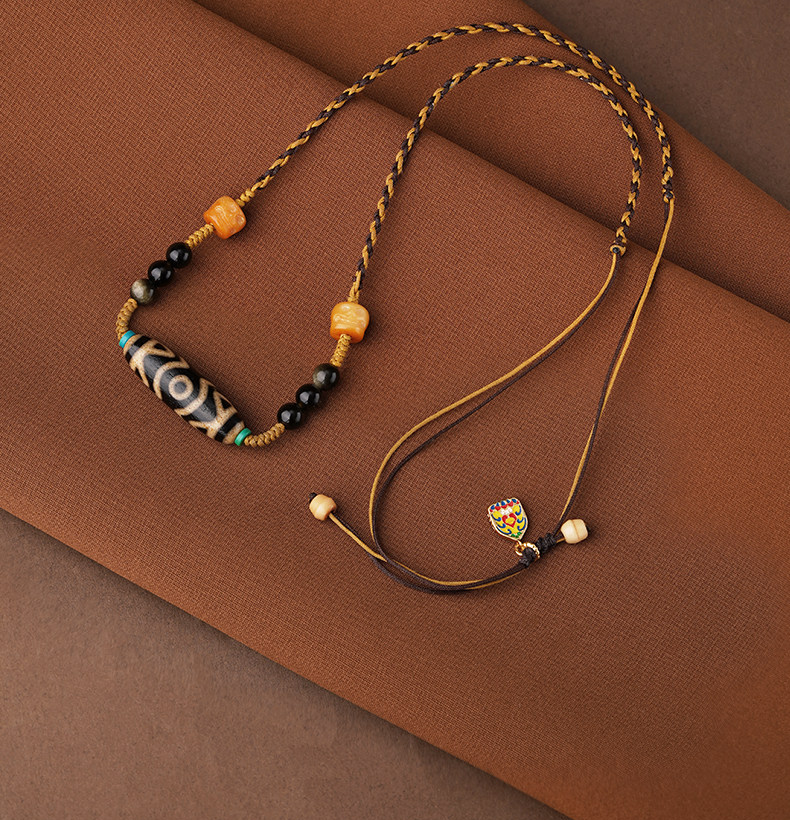 Inner Compass necklace on rust-colored fabric, showing its elongated eye-pattern bead, braided cord, pale tail beads and multicolor charm.