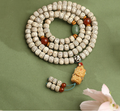 Cream speckled meditation beads coiled on a muted green surface, with red and green accents, a sculpted pendant and two beaded tails.