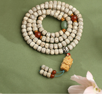 Cream speckled meditation beads coiled on a muted green surface, with red and green accents, a sculpted pendant and two beaded tails.