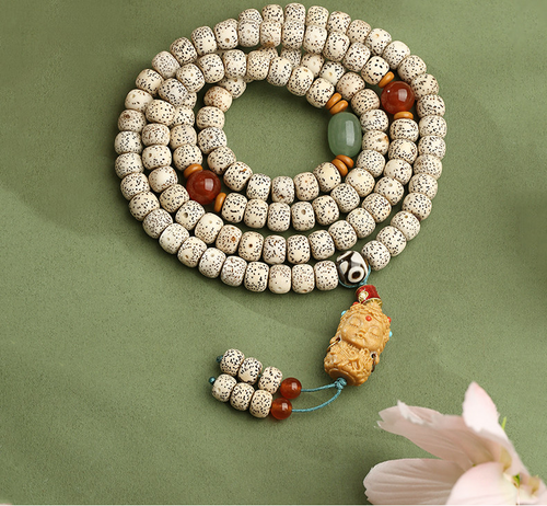 Cream speckled meditation beads coiled on a muted green surface, with red and green accents, a sculpted pendant and two beaded tails.