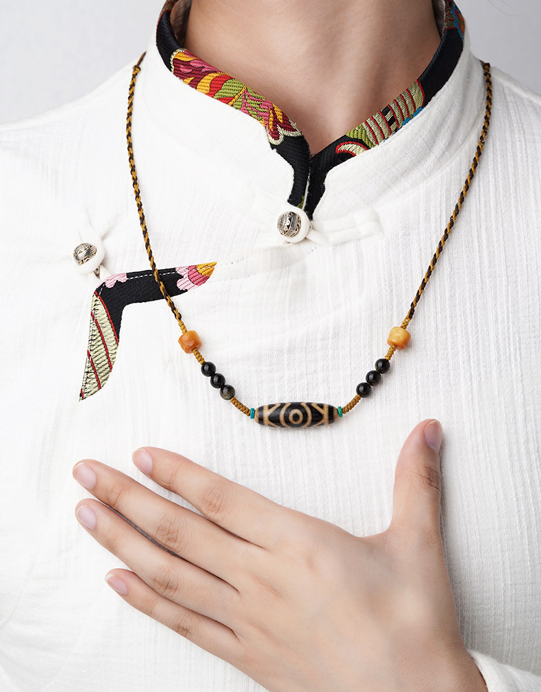 Eye-pattern cord necklace worn over a white collared top, with black accents and a mustard-and-black braided cord.