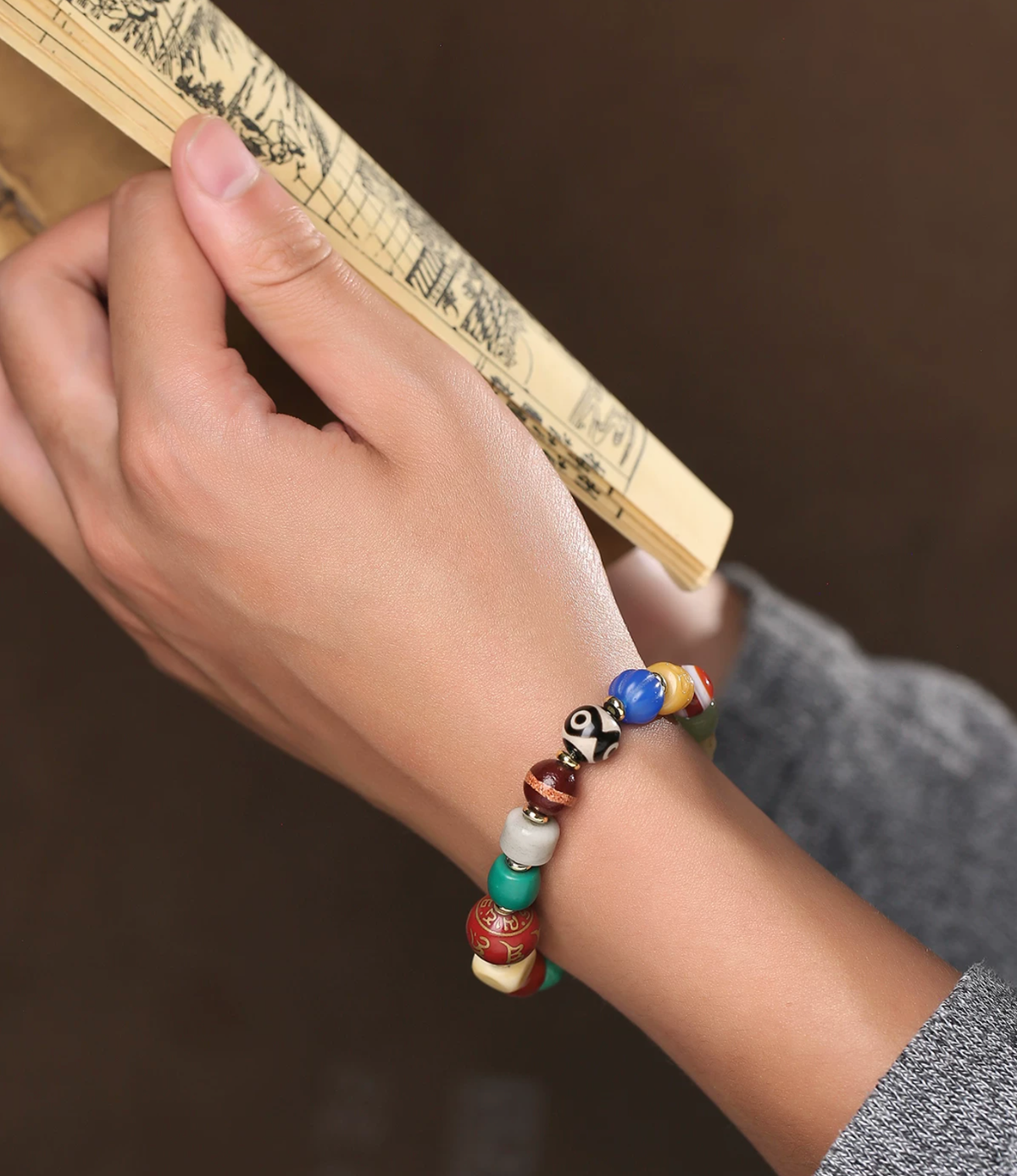 Multicolor bead bracelet on a wrist beside a folding fan, with glossy colored beads, sculpted details and gold-tone spacers.